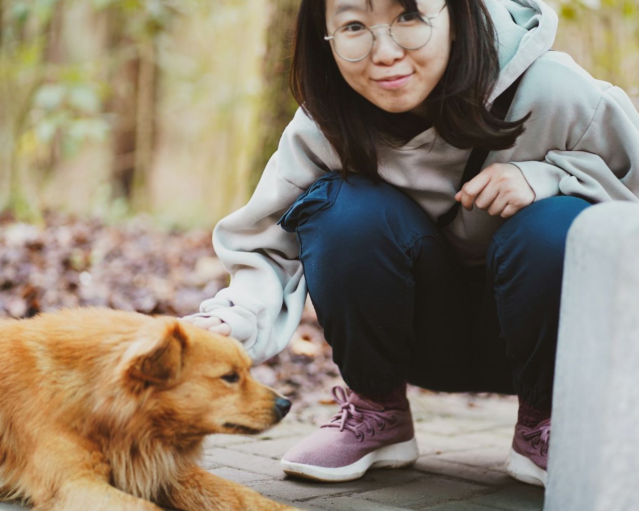 A woman crouching beside a golden dog on an autumn path, smiling at the camera